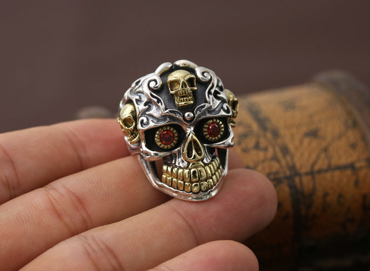S925 sterling silver skull ring in palm showing red zircon eye stones, brass crown detail, and oxidized finish